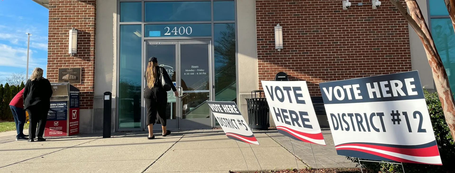 The front of a building with signage that reads 'Vote here', 'District #5' and 'District #12'. One person enters the building, whilst two others gather around a box that reads 'Official election ballot drop box'.