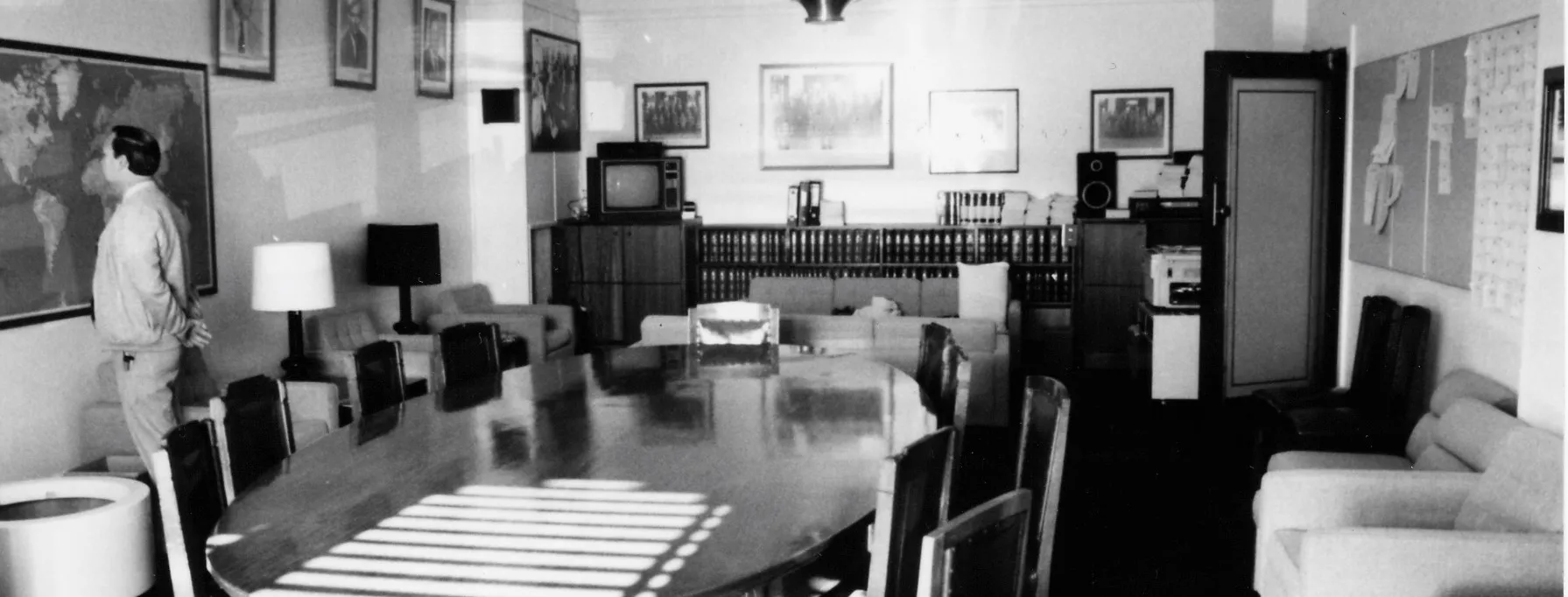 A large oval meeting table and chairs dominate the centre of the room. Easy chairs with side tables and lamps are arrayed along the sides and two sofas are arranged in a seating area towards the back of the room. A sideboard with books, a television and sound system stretches along the back wall. There is a framed map on one wall and a notice board on the opposite. Portraits of leaders and formal and informal photographs of the party crowd the walls