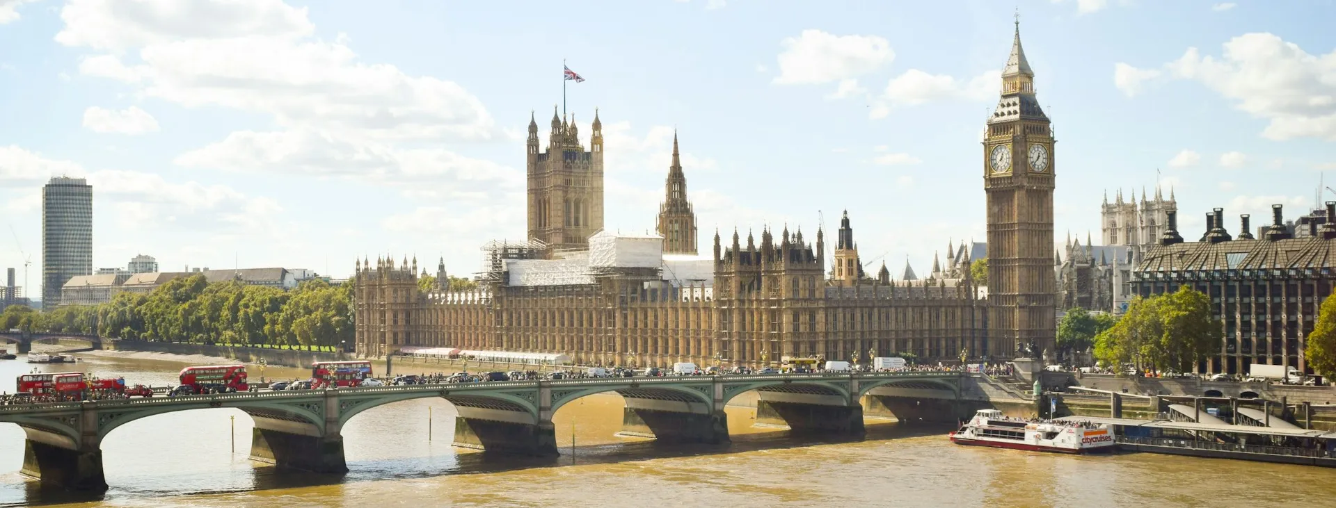 Big Ben and the Palace of Westminster, as viewed from across the river, with red double-decker buses and pedestrians crossing the bridge nearby.