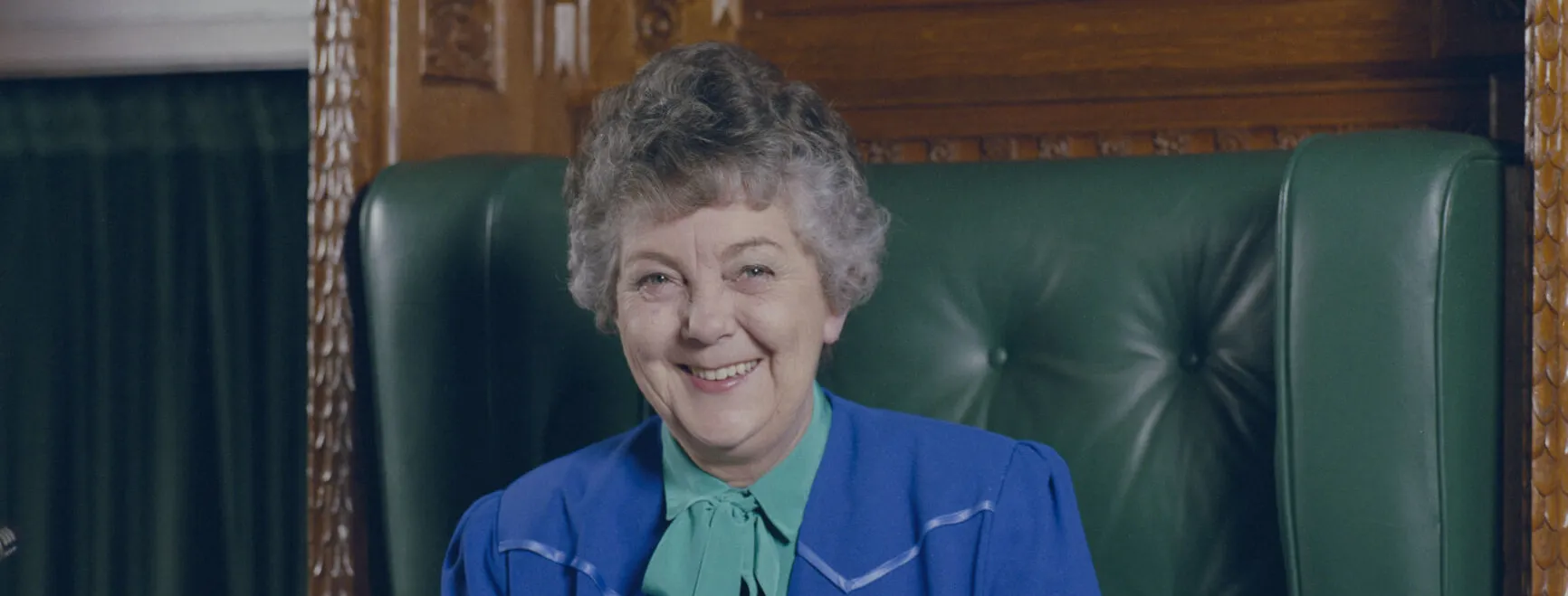 A colour photograph of Joan Child wearing a blue suit sitting on the green leather and timber Speaker's Chair.