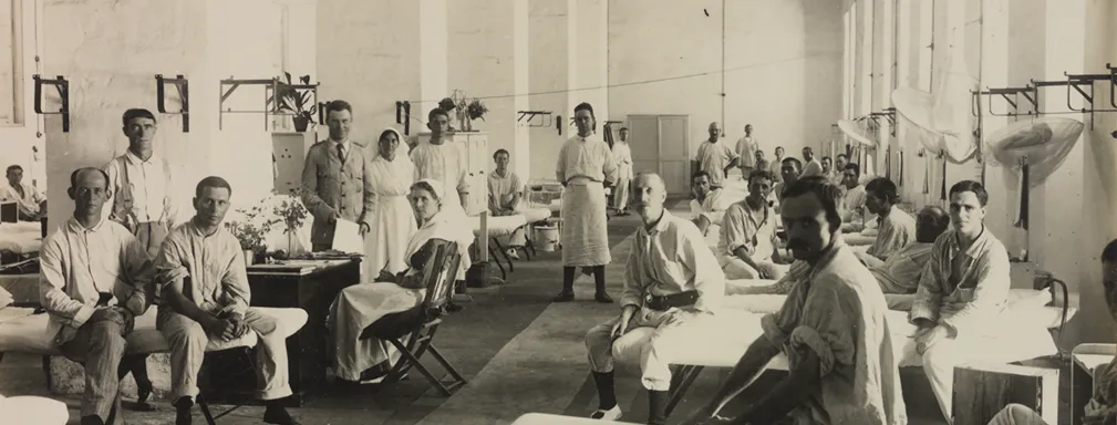 A surgical ward in Egypt during WWII with rows of beds with men looking at the camera. Earle Page is standing holding a piece of paper, next to a nurse. 