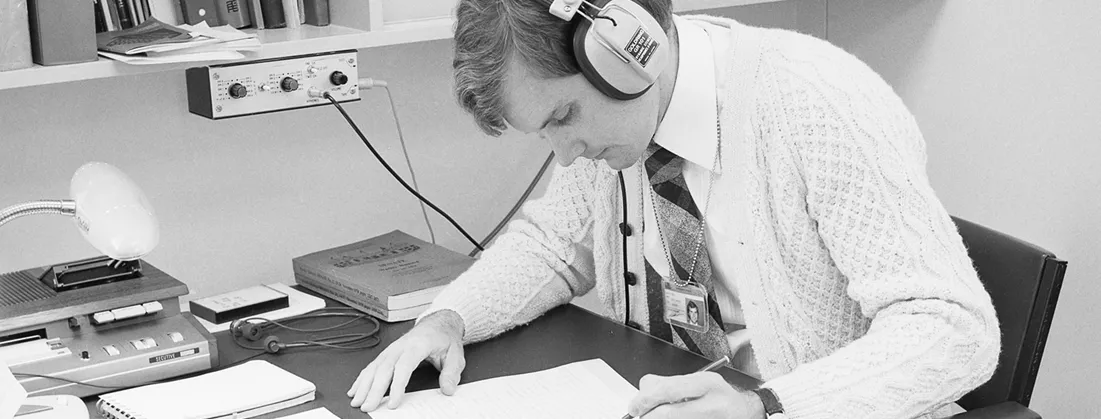 A man with headphones on is writing on a sheet of paper. He is in an office surrounded by papers.