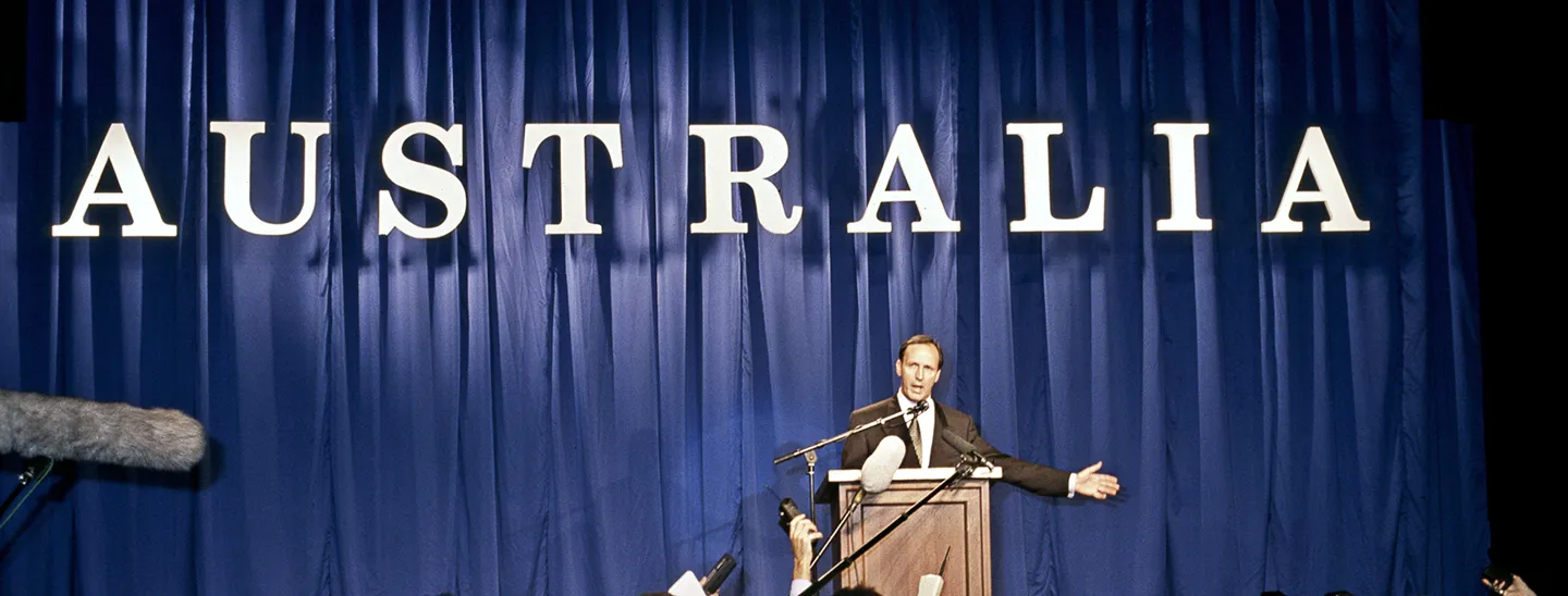 Paul Keating stands at a lecturn with the word 'Australia' behind him. A crows 