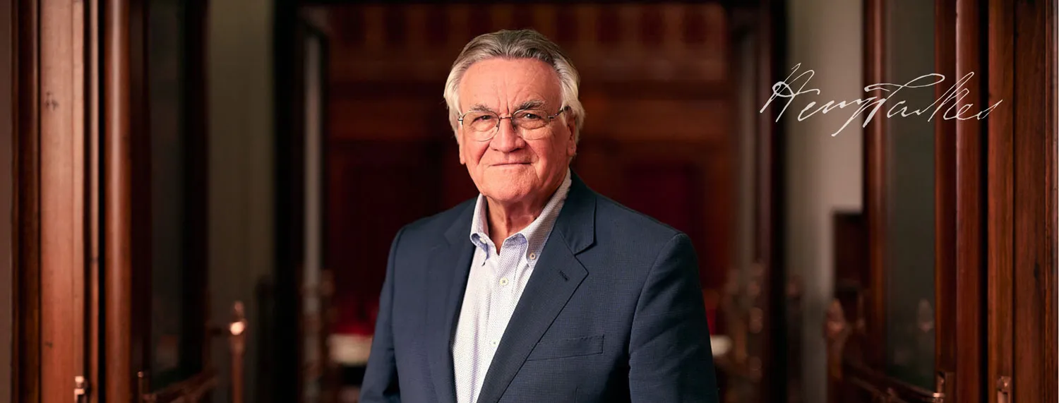 Barrie Cassidy wears a blue suit with light blue shirt. He is standing in Old Parliament House in an ornate wooden doorway.