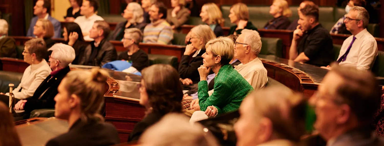 An audience sits on green leather benches in the House of Representatives at Old Parliament House. The room has wood panelled walls.