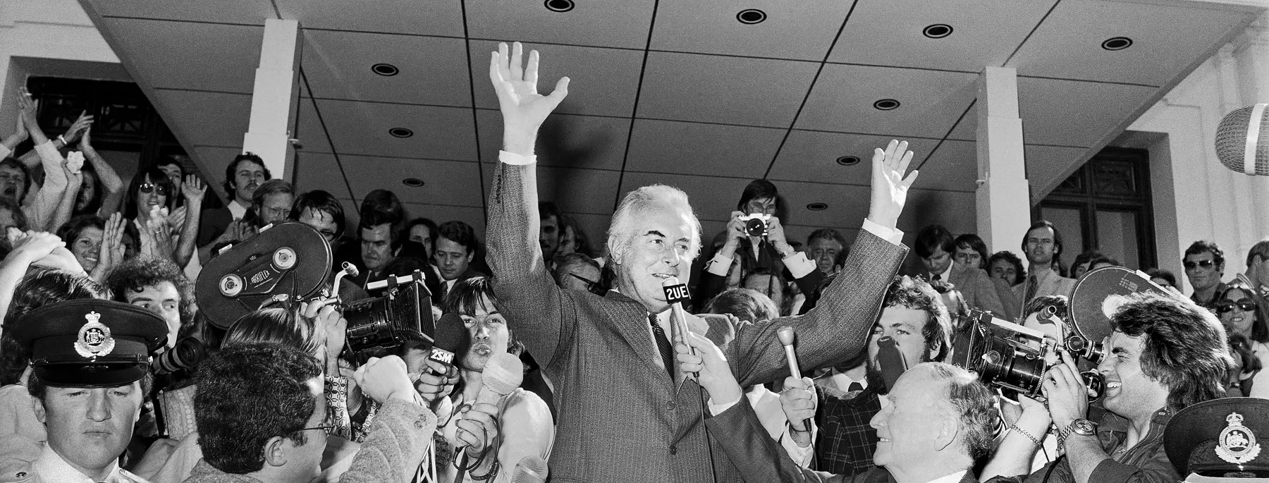 Gough Whitlam stands with his hands in the air amidst a crowd of reporters with microphones and cameras on the steps of Old Parliament House on November 11 1975.