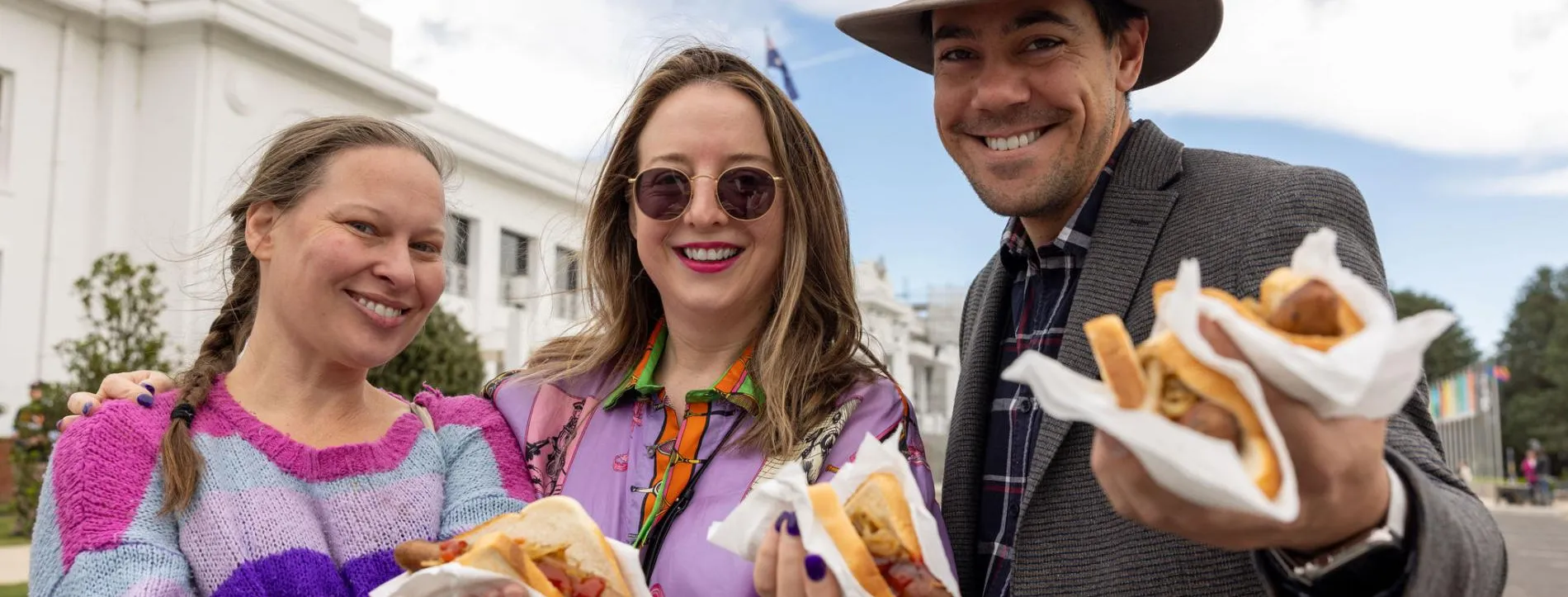 Two women wearing brightly coloured purple tops and a man in a jacket and hat stand in front of Old Parliament House holding sausage sandwiches.