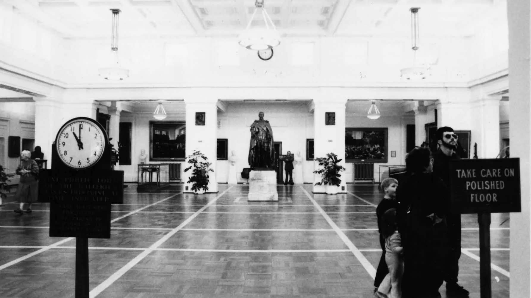 This black and white photograph of King’s Hall looks along the length of the room to the slightly larger than life bronze statue of King George the Fifth on a marble pedestal. King’s Hall is a large white room with a vaulted ceiling, celerestory windows, colonnades and a polished parquet floor.  The walls and columns feature artworks and portraits. You can see people wandering through the space including a family with young children and an older lady. In the foreground are two signs.