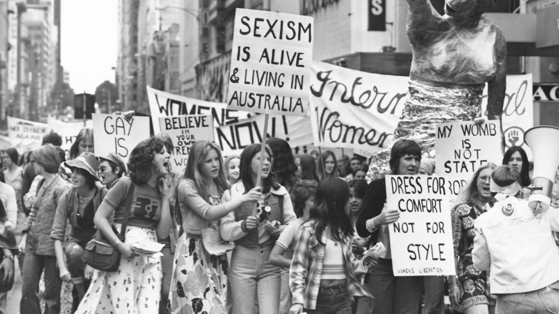 A black and white photo of a group of women holding protest signs in 1975. Signs say 'sexism is alive and well in Australia' and 'dress for comfort not style'.