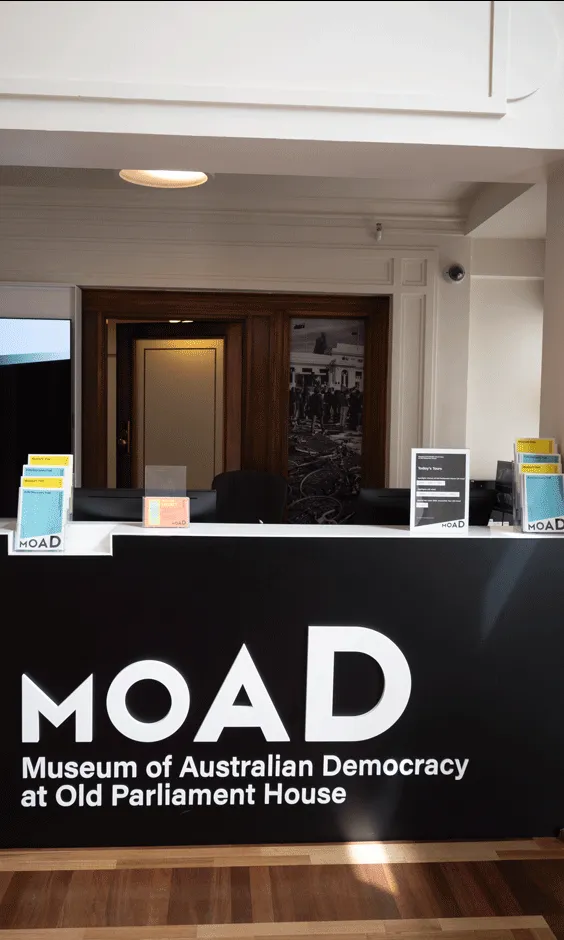 A front desk with pamphlets on top of it and a large black sign with the words 'MoAD Museum of Australian Democracy at Old Parliament House.'