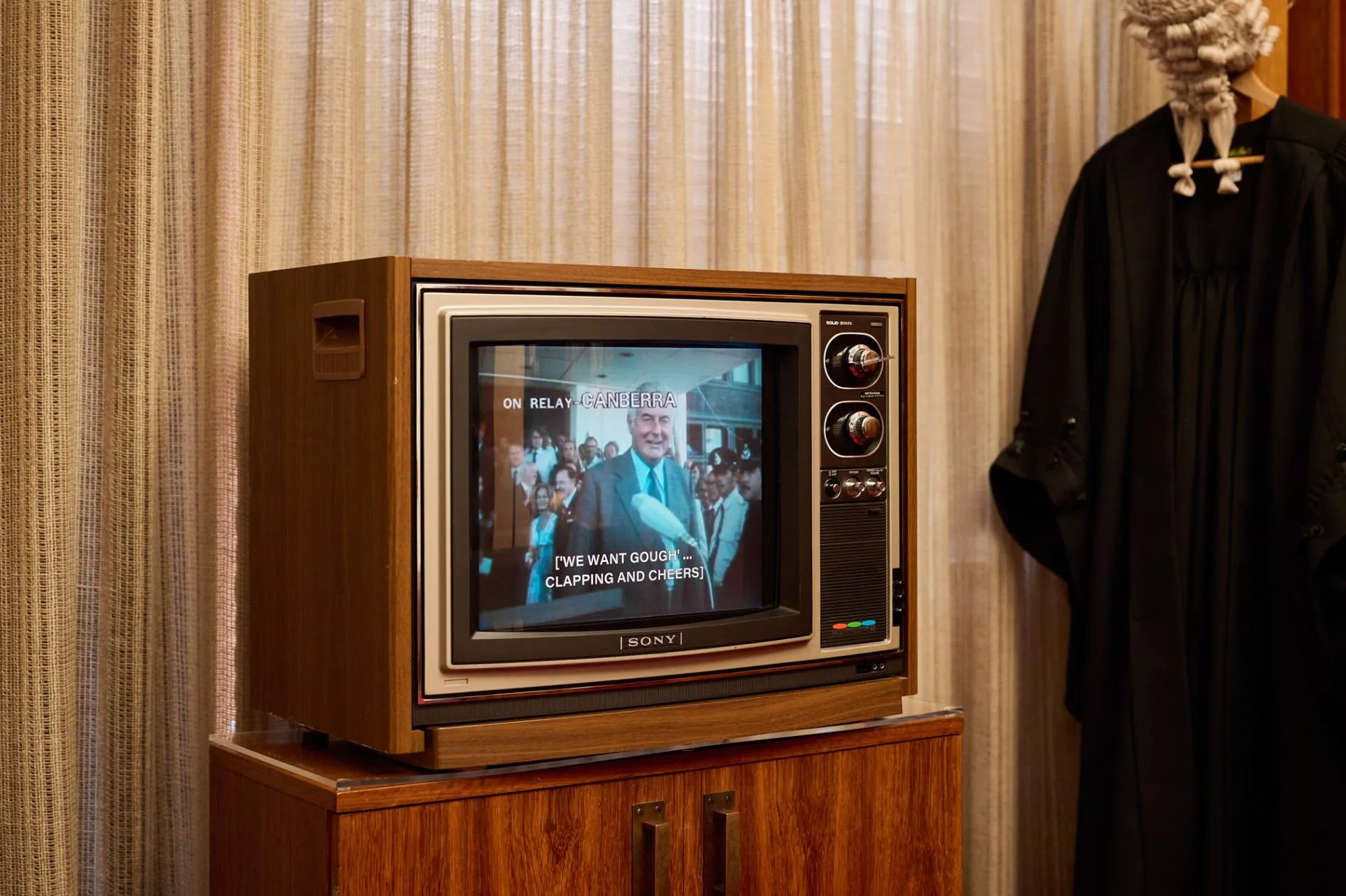 A wood-framed TV with an image of Gough Whitlam sits on a wood cabinet. A black gown and white wig hang next to the TV.