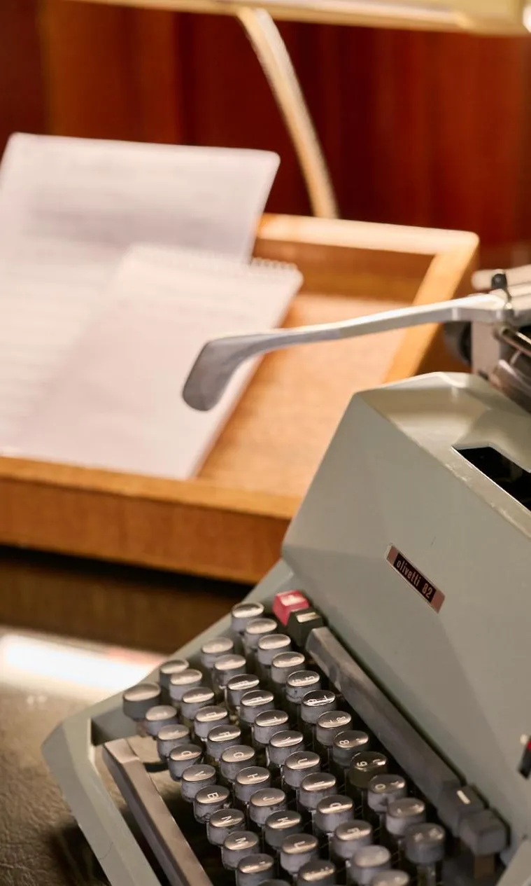 An old-fashioned grey typewriter sits on a desk next to a wooden try with documents in it.