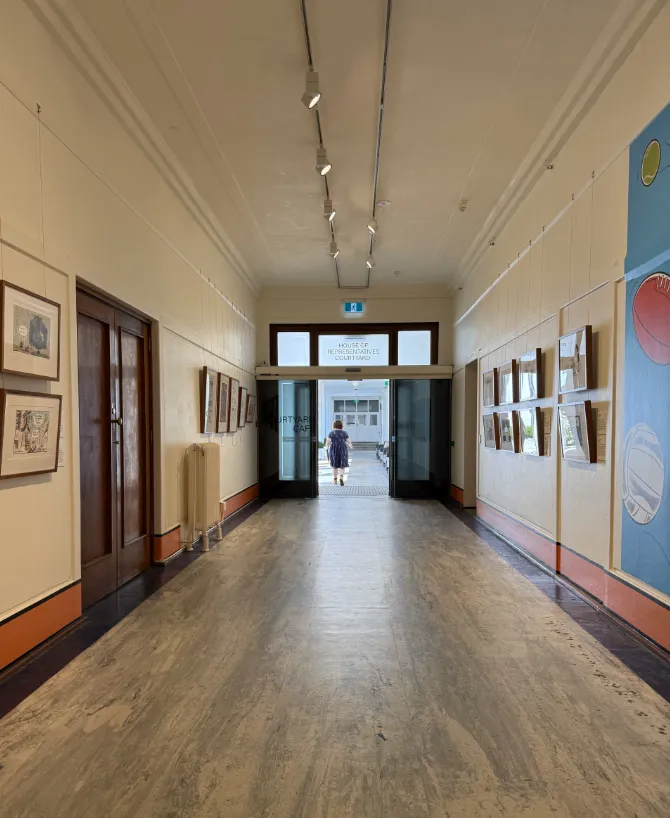 A hallway with cartoons hung on the walls. At the end of the walkway a glass door is open leading to a courtyard. 