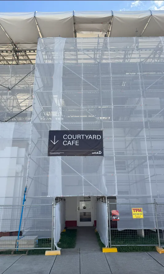 White transparent scaffolding covers the entrance to a building. There is a large black sign with the words Courtyard Cafe and an arrow pointing down to the doorway in the scaffolding.