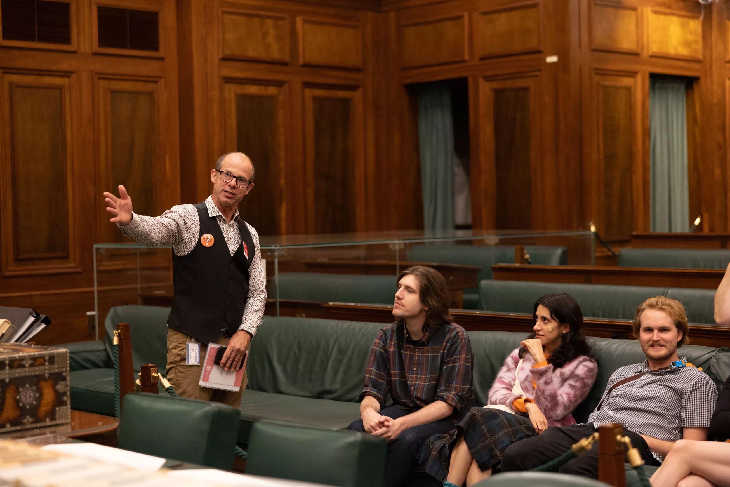 A volunteer has his hand out as he describes something to 3 people sitting in the green leather seats of the House of Representatives Chamber. 