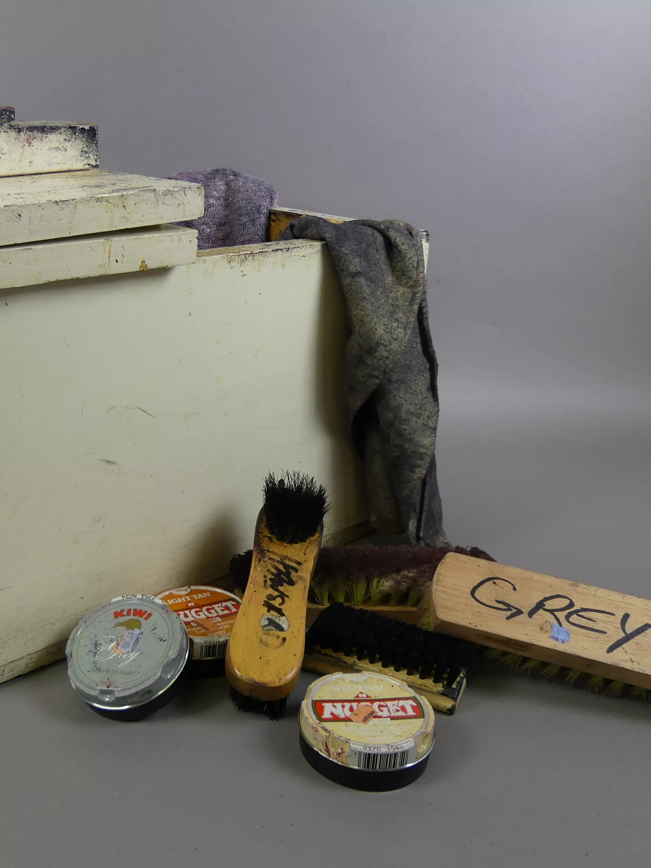 A solid wooden box containing dirty cloths, four shoe brushes and three tins of shoe polish.