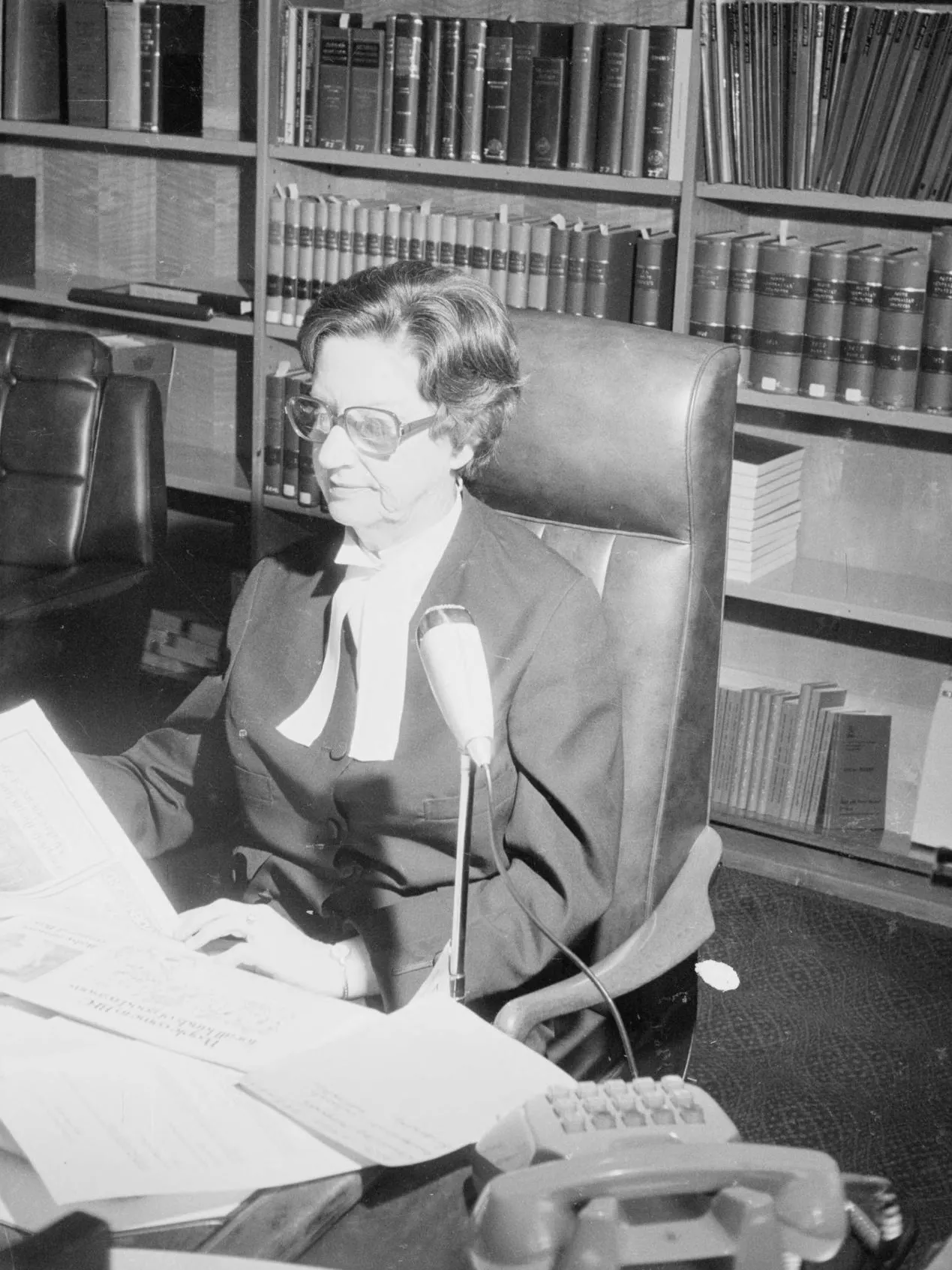 A woman, Justice Roma Mitchell sits at a desk covered in papers, in front of a bookshelf filled with bound volumes.