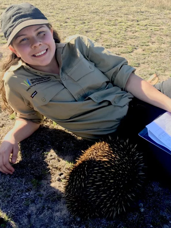 Dhani Gilbert lies on the ground, looking at the camera, with an echidna close to her