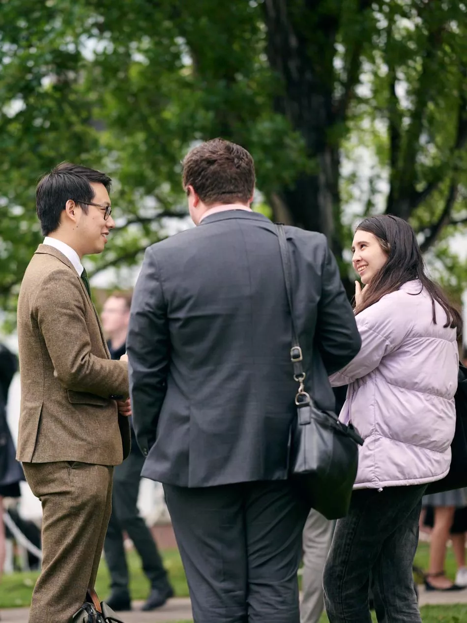 A group of three people talking together in the foreground, with many other people in the background, under a large tree in a courtyard.