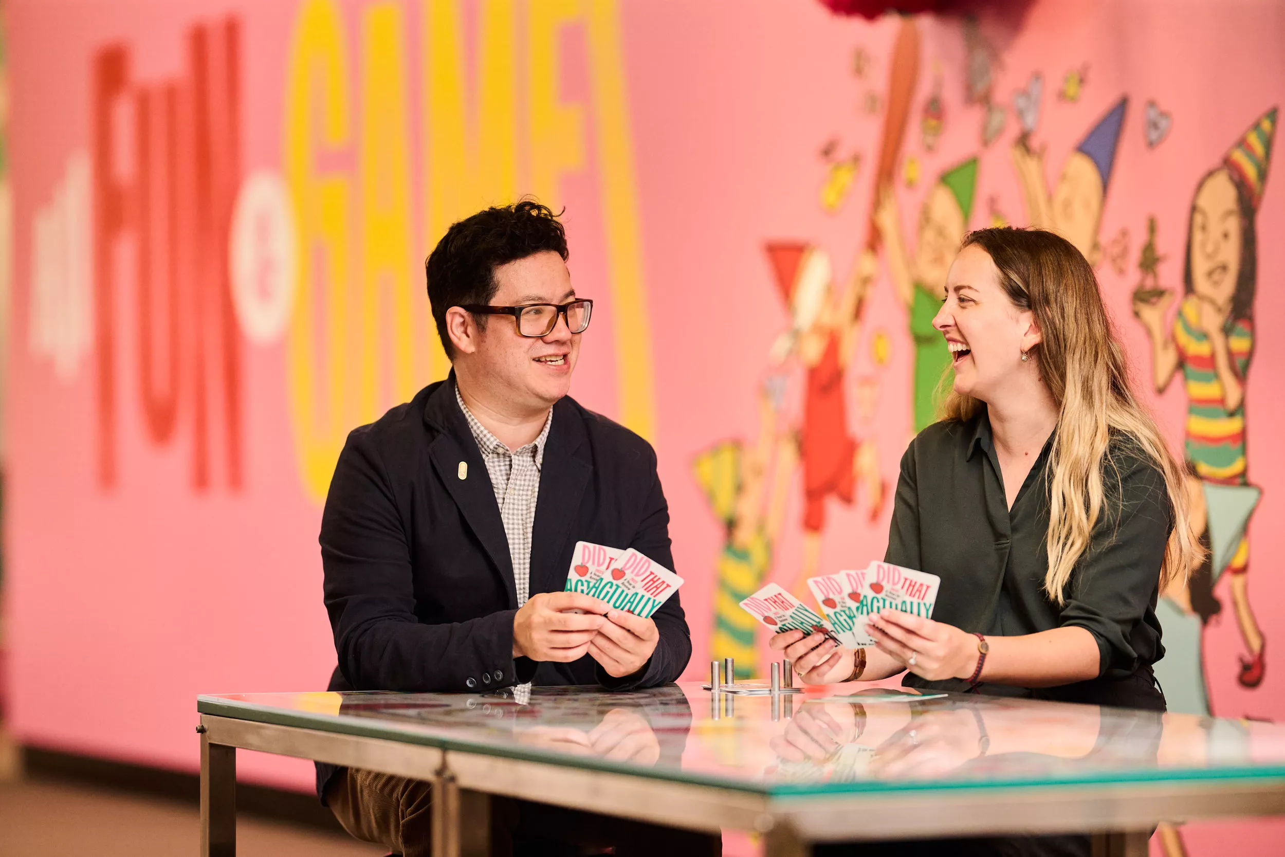 Two people seated at a table playing a card game in front of a brightly decorated wall.