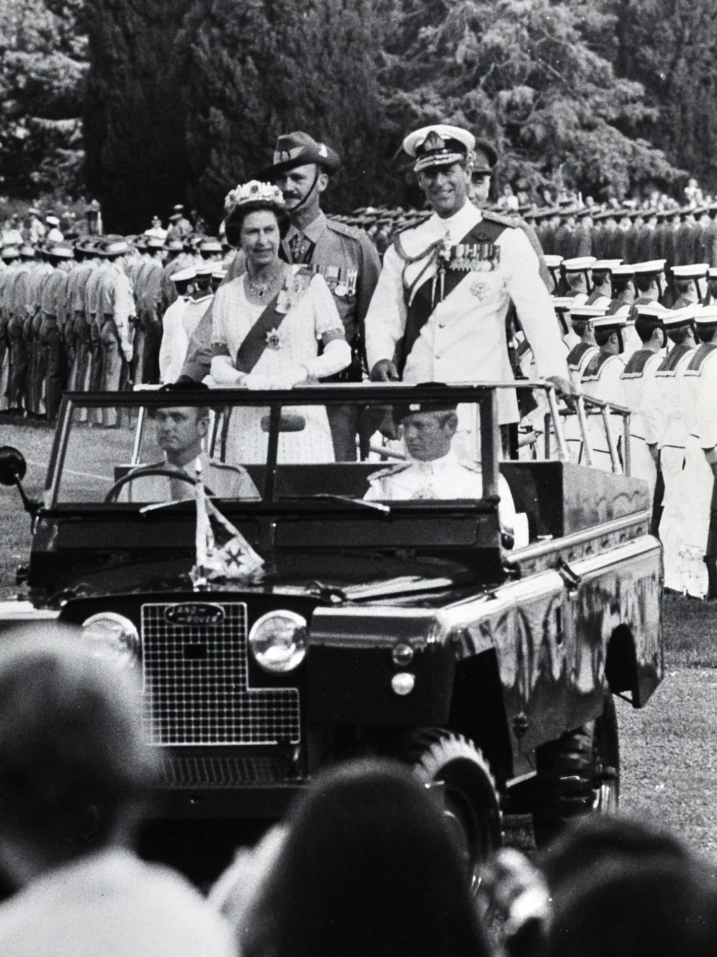 Queen Elizabeth II and Prince Philip stand on the back of an open-topped car, passing troops standing in formation.