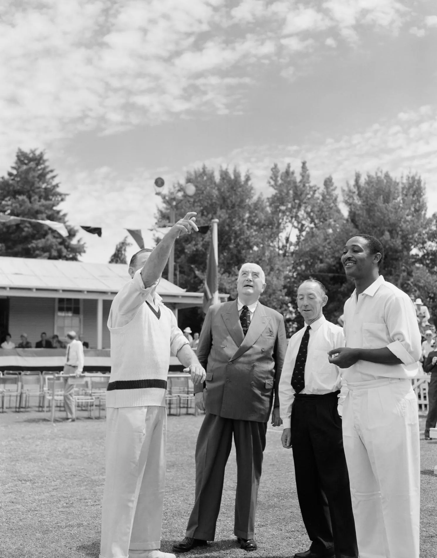 Prime Minister Menzies and the captains of the Prime Minister's Eleven and the West Indies cricket teams both wearing cricket whites toss coins on a cricket oval.