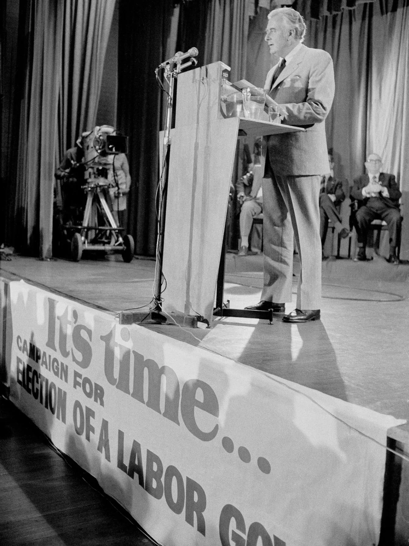 Gough Whitlam stands on a stage at a podium with a microphone launching Labor's election campaign. There is a large sign on the front of the stage which says, 'It's time... Campaign for election of a labor gov...'