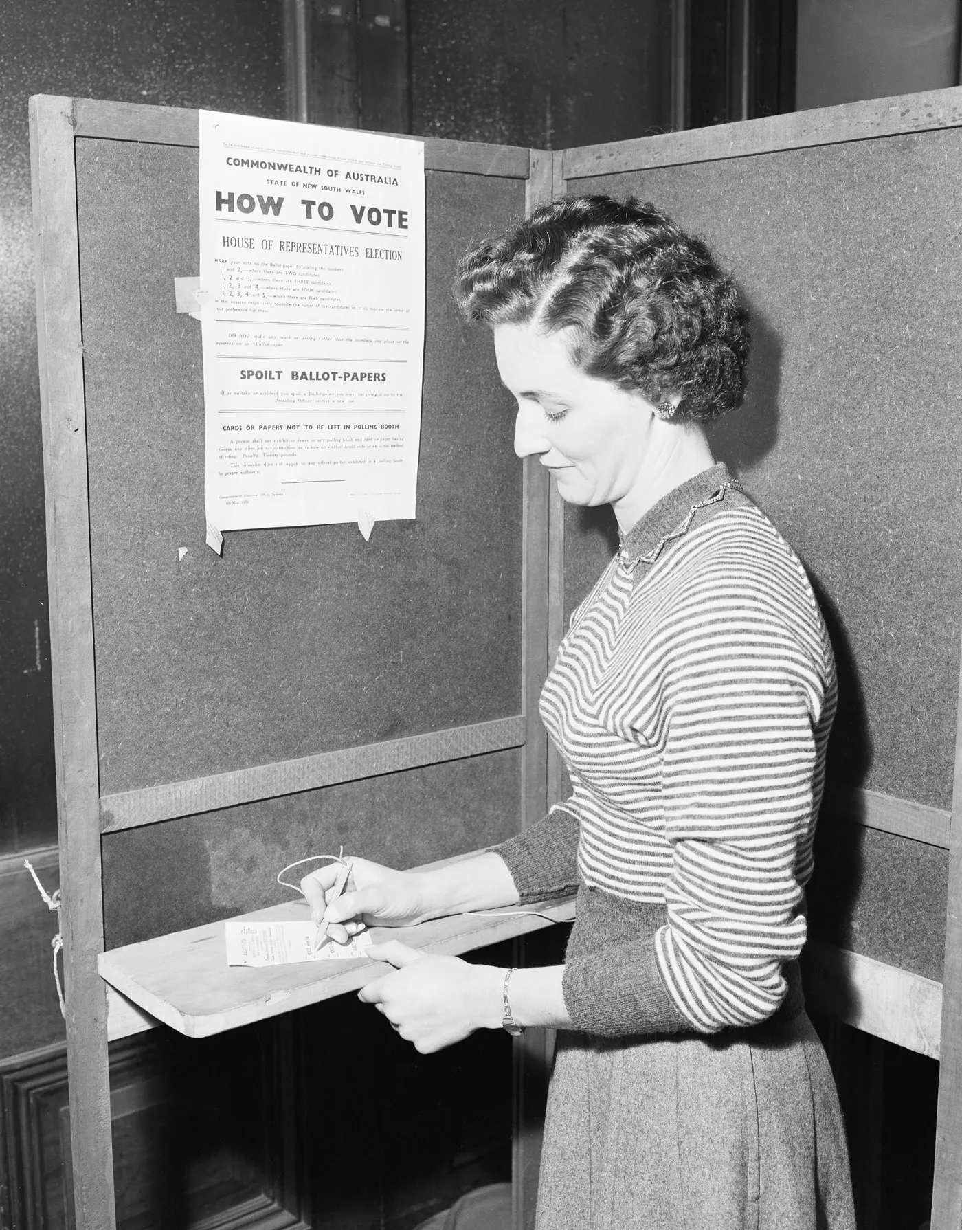 A woman in a striped sweater stands in a polling booth holding a pencil above a voting card. A 'How to vote' instruction poster is pinned to the booth wall.