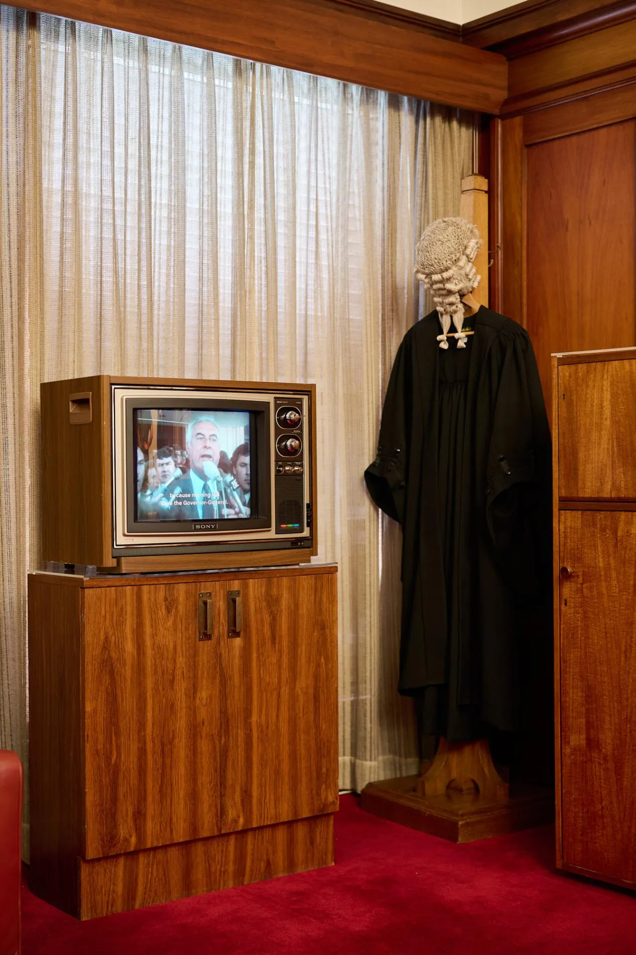 In a room in Old Parliament House a wooden TV with an image of Gough Whitlam is on a wooden cabinet next to a stand with a black gown and white wig. There is red carpet on the floor.