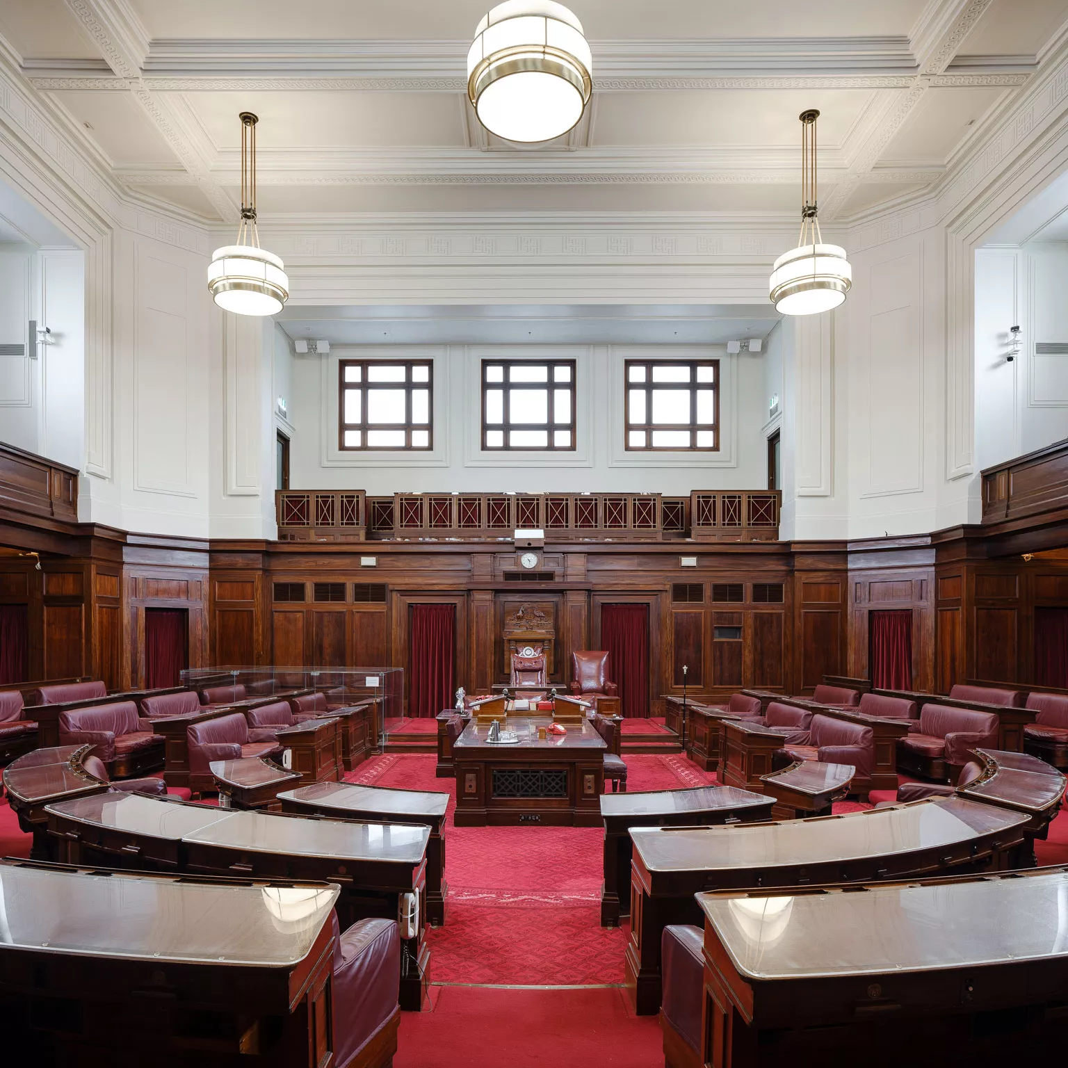 A view into the Senate Chamber looking straight into the room with benches in a U-shape on either side of the centre table. The carpet is a rich red and the seats are red leather. Art deco lights hang from the ceiling and wood timber panels surround the room. 