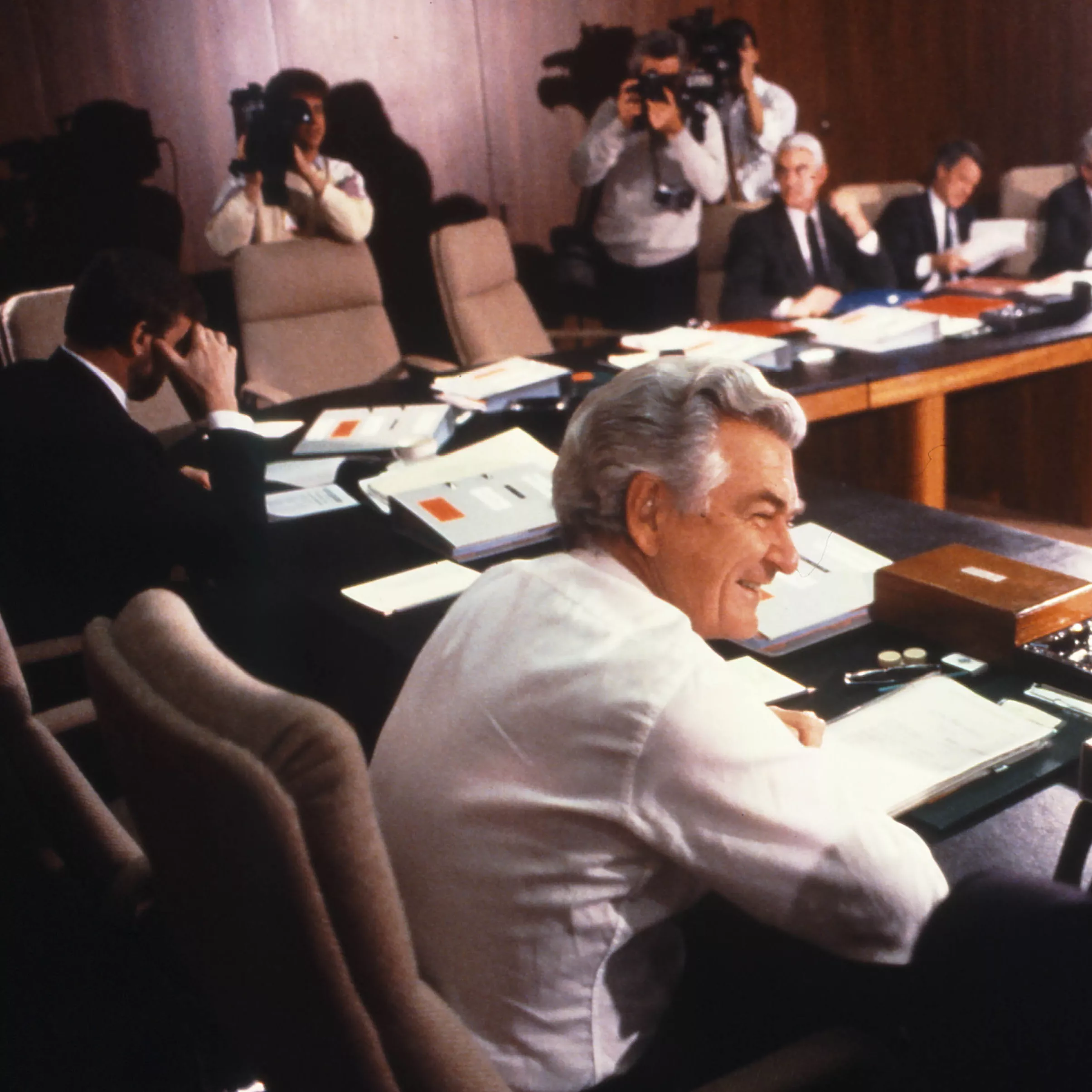 This colour photograph is taken from behind Deputy Prime Minister Lionel Bowen who is seated beside Prime Minister Bob Hawke. Both are dressed in standard business attire.  Other members of Cabinet can be seen sitting around the table although only half of the seats are filled. In the background are a photographer and two cameramen photographing and filming the cabinet meeting. 