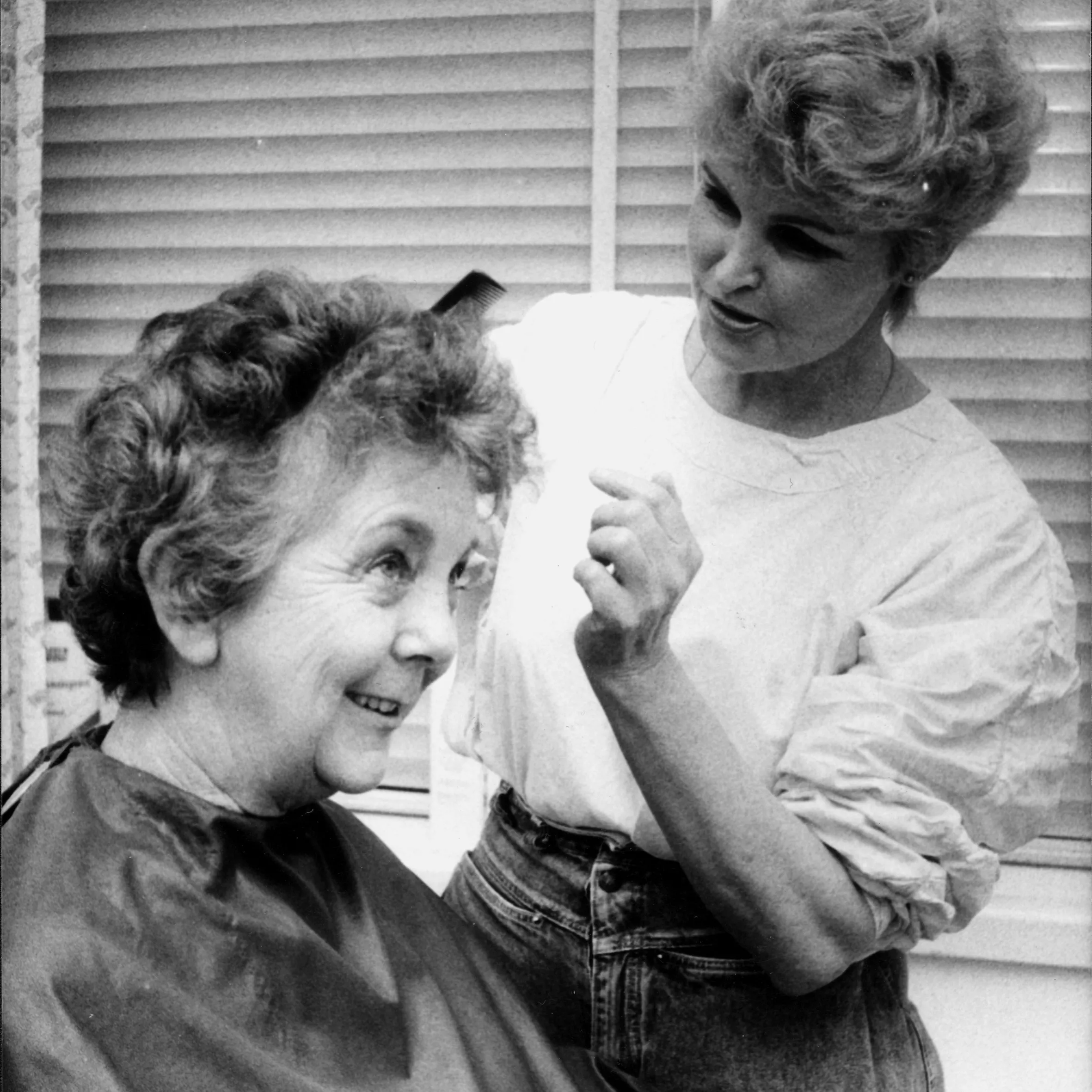 This black and white photograph depicts hairdresser Lizzie Scott, arranging Speaker Joan Child’s hair. Joan is sitting in the barber’s chair and is covered with a dark  cape while Lizzie stands to her left wearing a white shirt and jeans and holding a black comb.  