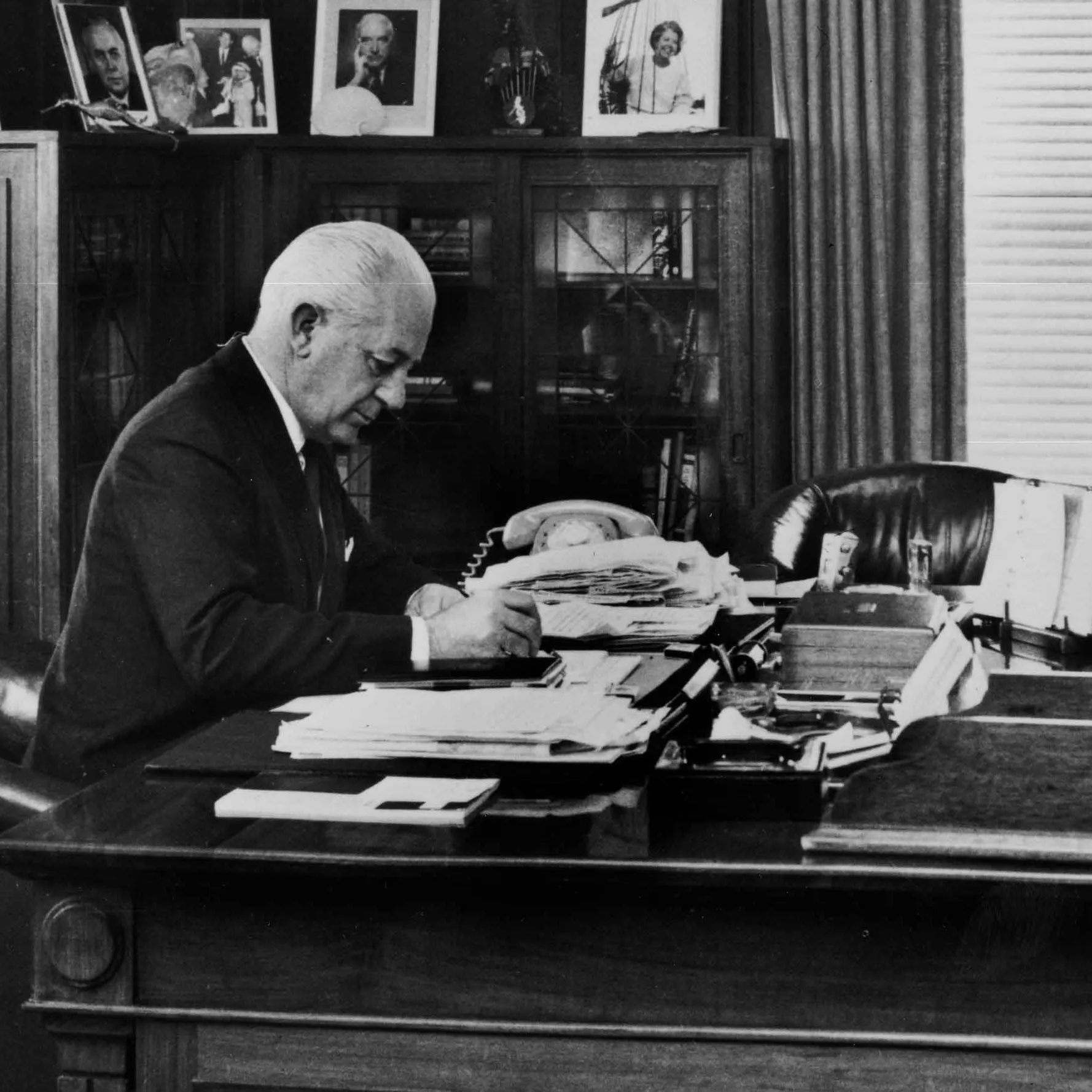 In this black and white photograph Prime Minister Harrold Holt sits at the timber desk made in 1926. Holt is wearing a dark suit, white shirt and tie and is leaning on the desk while he writes. The desk has fine carved panels with a geometric detailing on the corners. The surface of the desk is cluttered with stacks of documents and a telephone. On the glass-fronted cabinets in the background are a number of framed photographic portraits.  