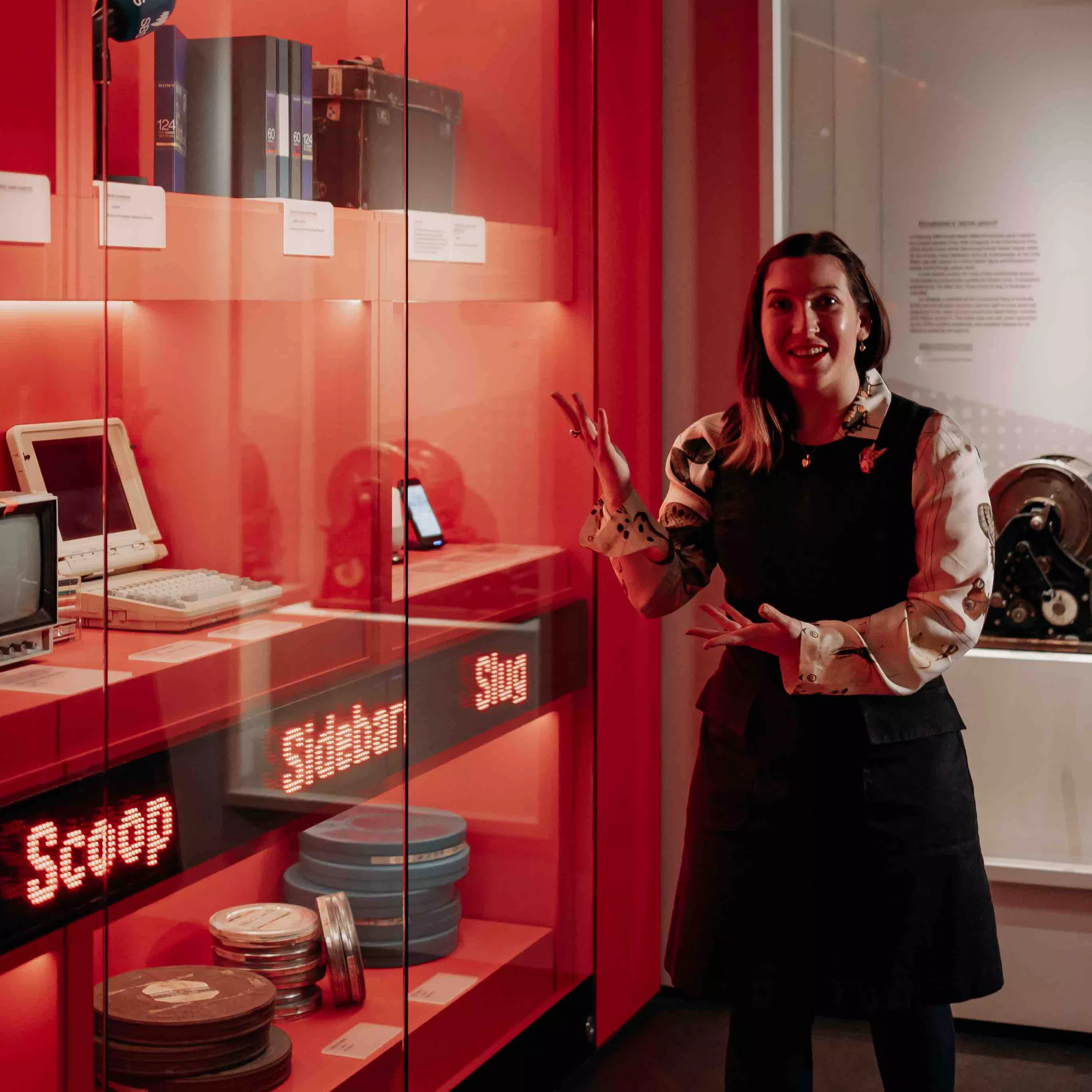 A MoAD educator stands in front of the Truth, Power and a Free Press exhibition wall featuring red walls with different types of media technology behind the glass case. 