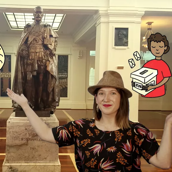 A MoAD educator wearing a hat, gestures with her hands in front of a green screen that features King's Hall in Old Parliament House superimposed with cartoon characters. 