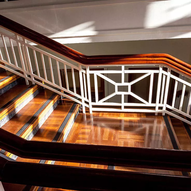 A close up of a timber staircase with dappled light. The railings feature white posts with an art-deco style design.