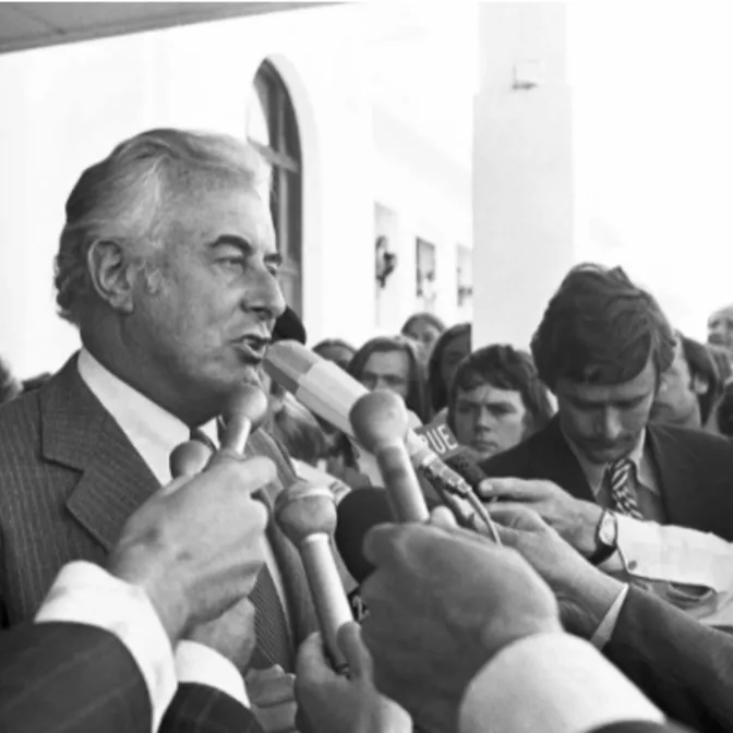 Black and white photograph of Gough Whitlam speaking to a large group of people holding microphones to his face, in front of parliament house immediately following the Dismissal.