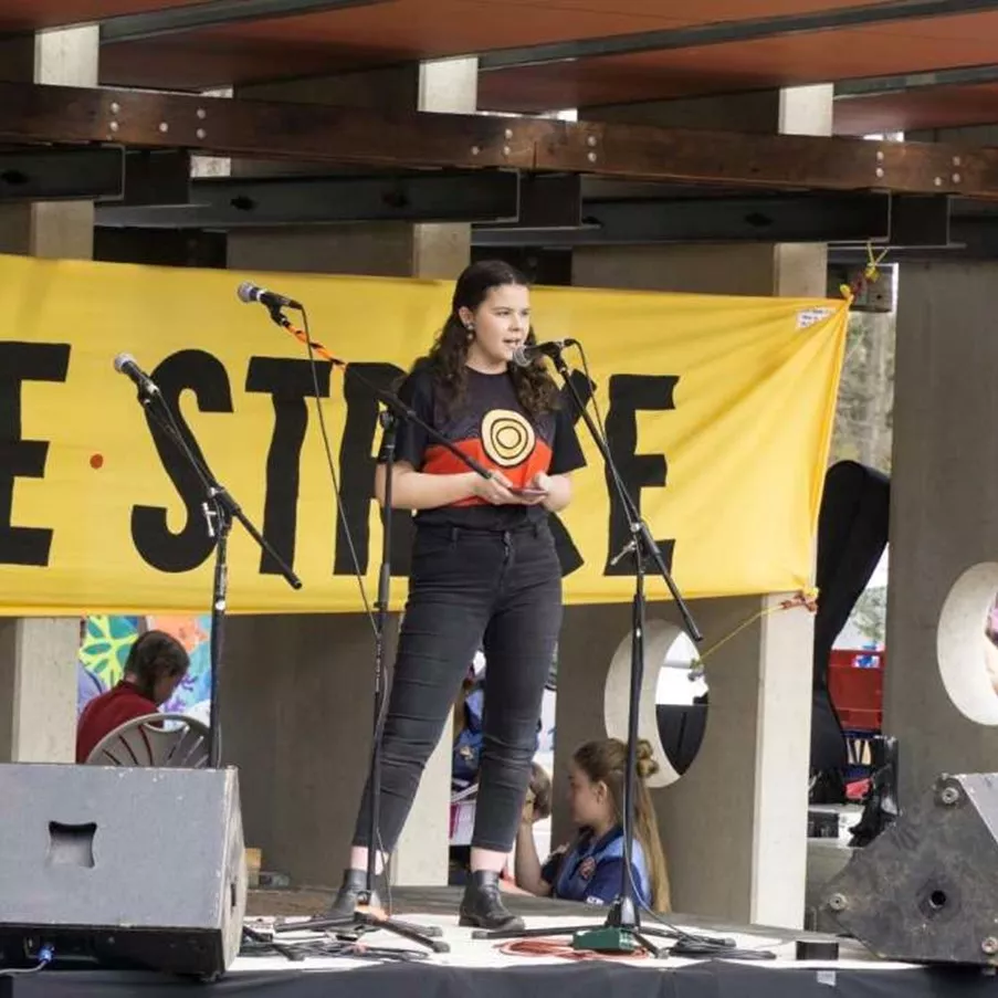 Dhani Gilbert speaks into a microphone, on stage, for a school strike for climate change protest