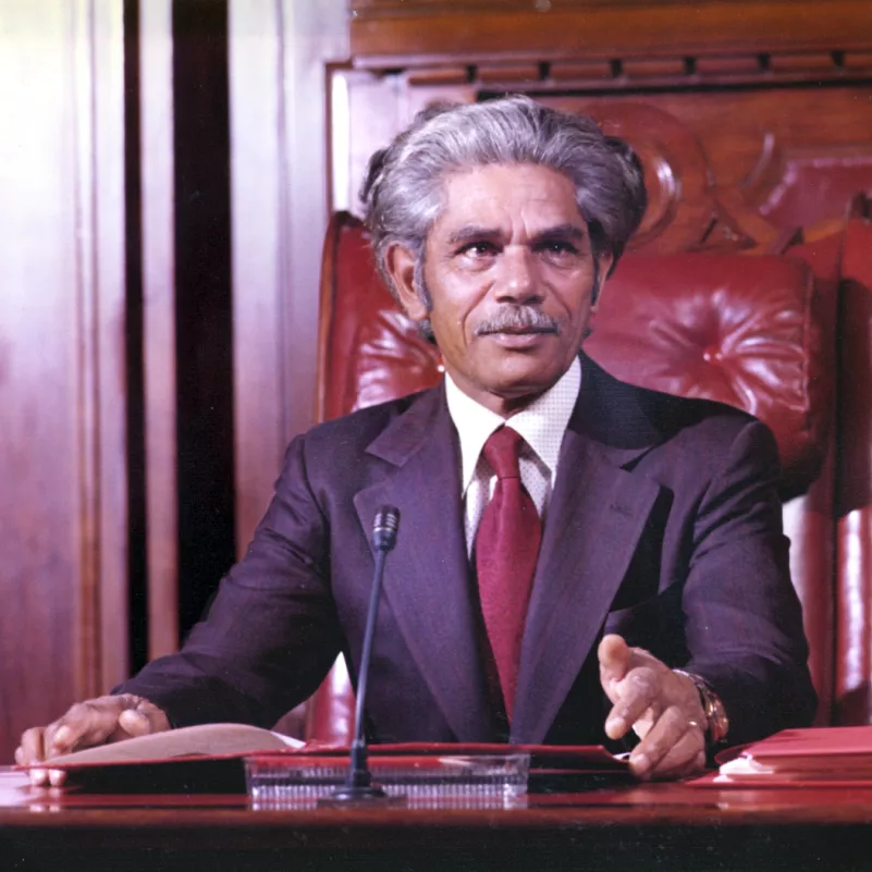 Neville Bonner seated in the Senate chamber, wearing a suit and red tie.