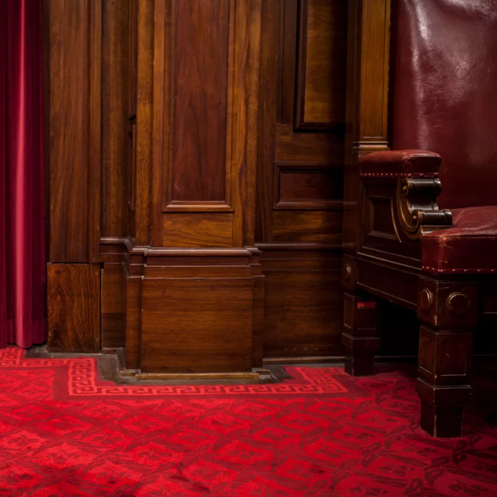 Close up of a red velvet curtin falling to a red carpeted floor with a timber furniture panel next to it.