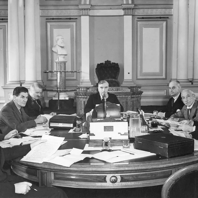 Nine men are gathered, seated around a large table covered with paper documents, with Robert Menzies seated at the head of the table.