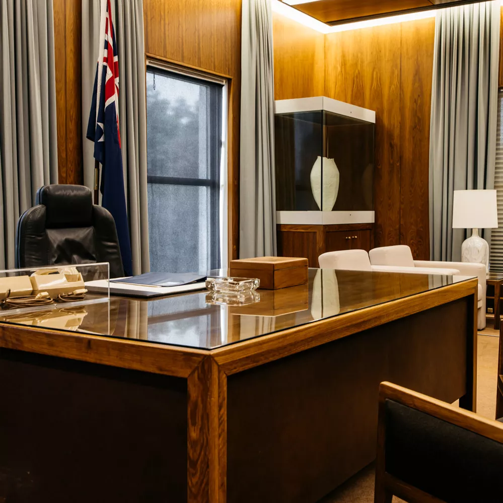 A colour photograph of the Prime Minister's Desk. Behind it is a comfortable office chair and an Australian flag on a pole. In front and to the side of the desk are armchairs.