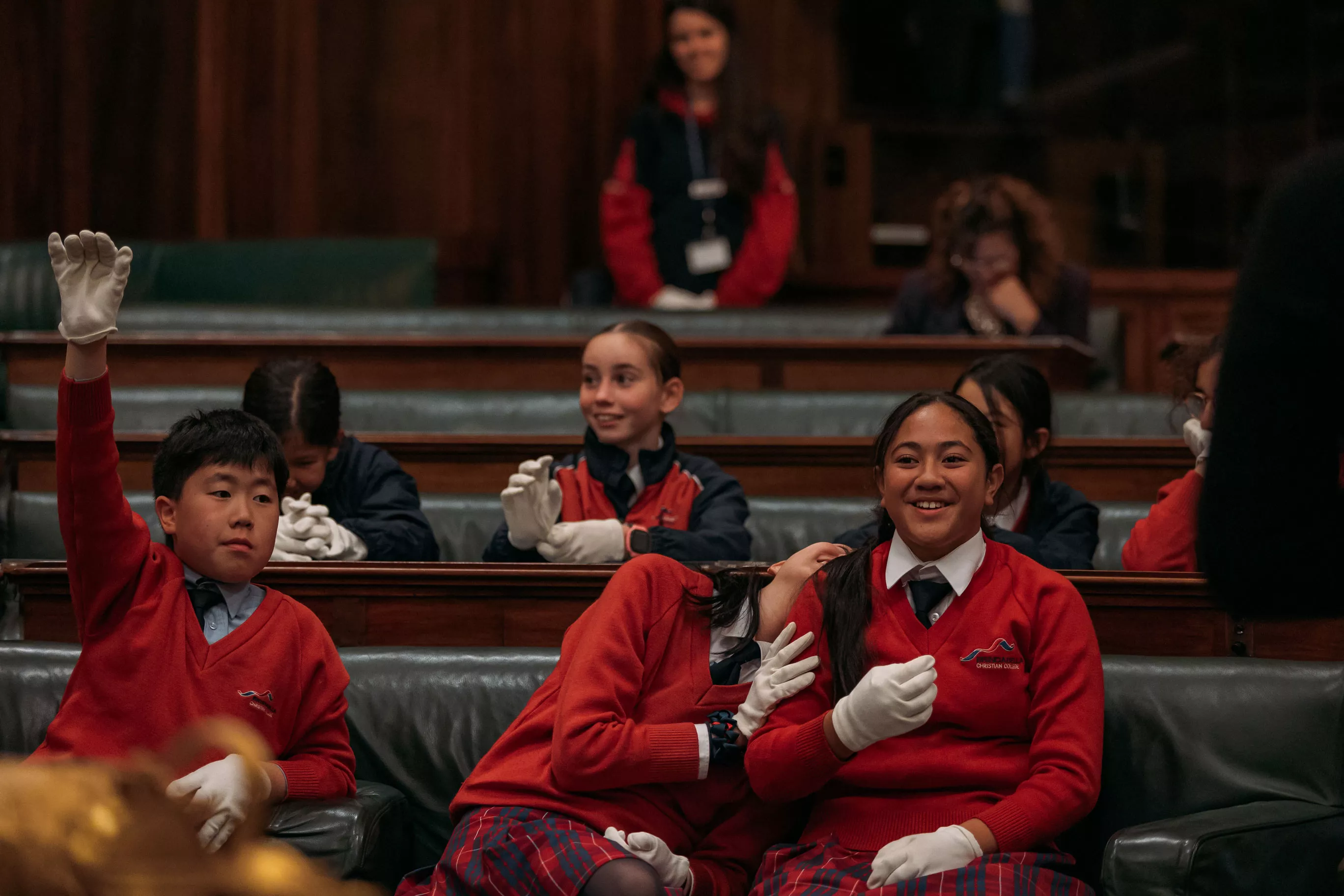 High school students in red jumpers sit on green leather seats in the House of Representatives Chamber. Two girls are laughing. 