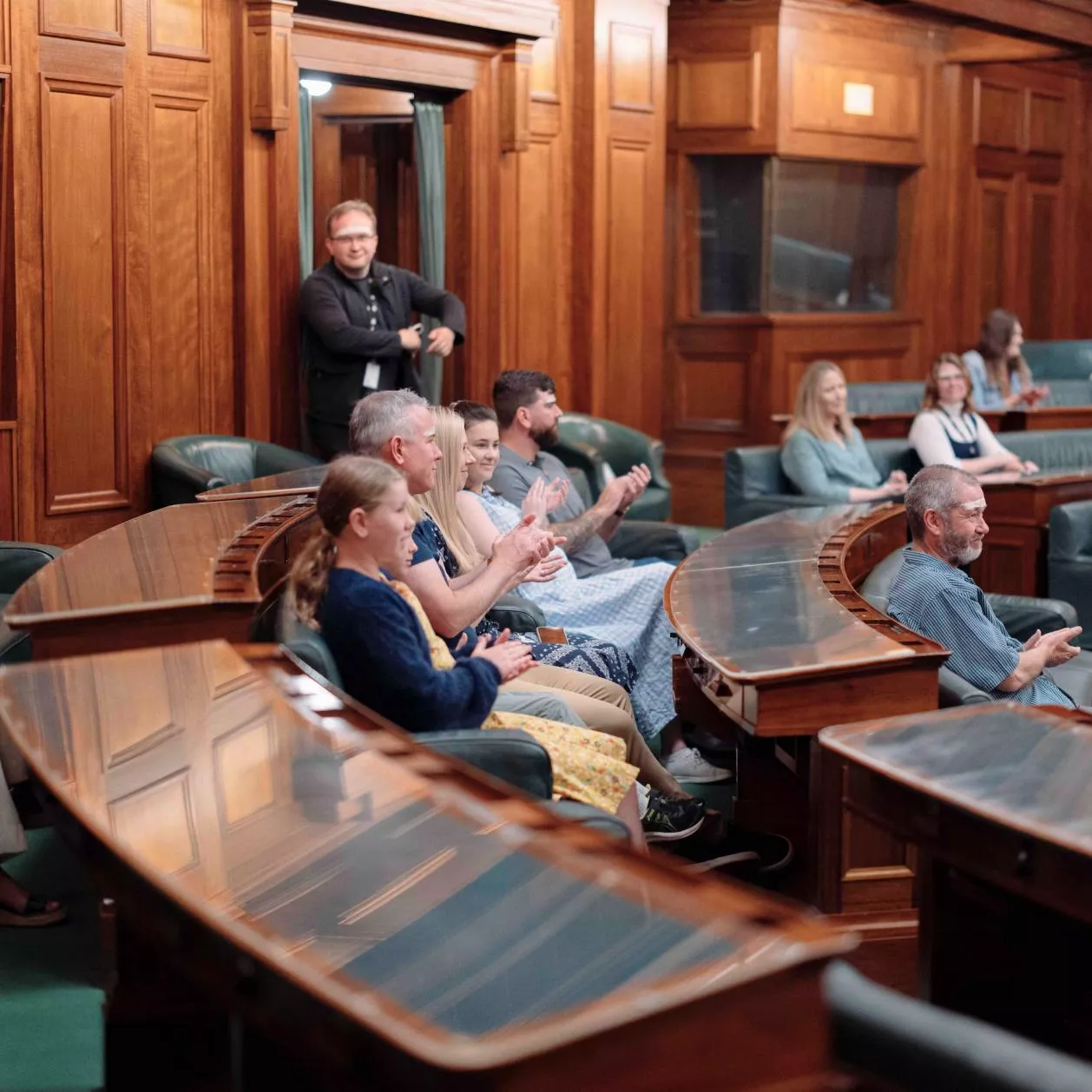 Visitors sit in green leather chairs with timber desks with glass coverings in a U-shape in the House of Representatives. 