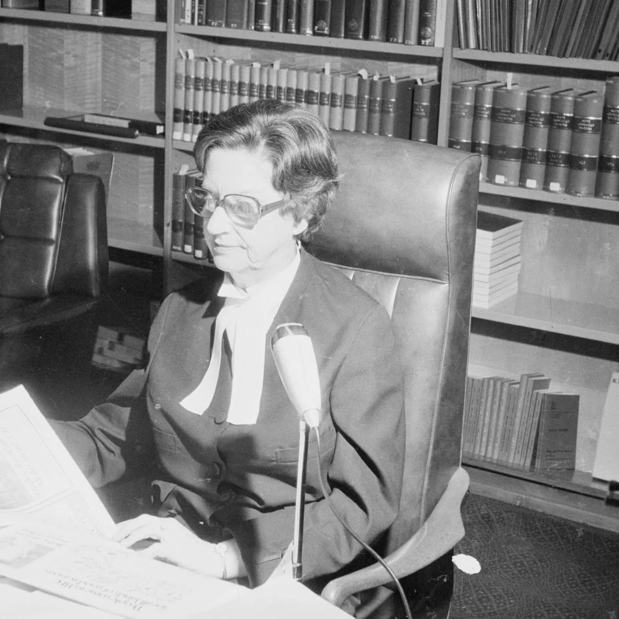 A woman, Justice Roma Mitchell sits at a desk covered in papers, in front of a bookshelf filled with bound volumes.