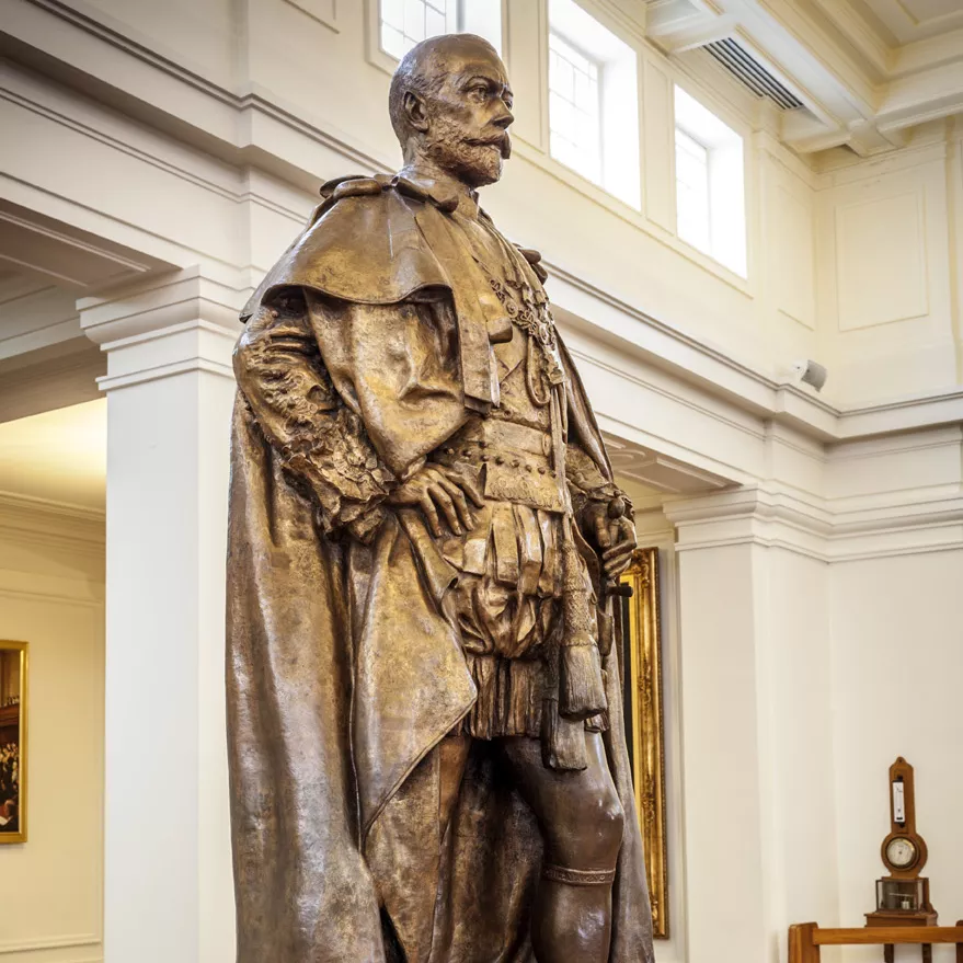 A bronze statue of a man with a cloak on a marble pillar in a room with timber floors.