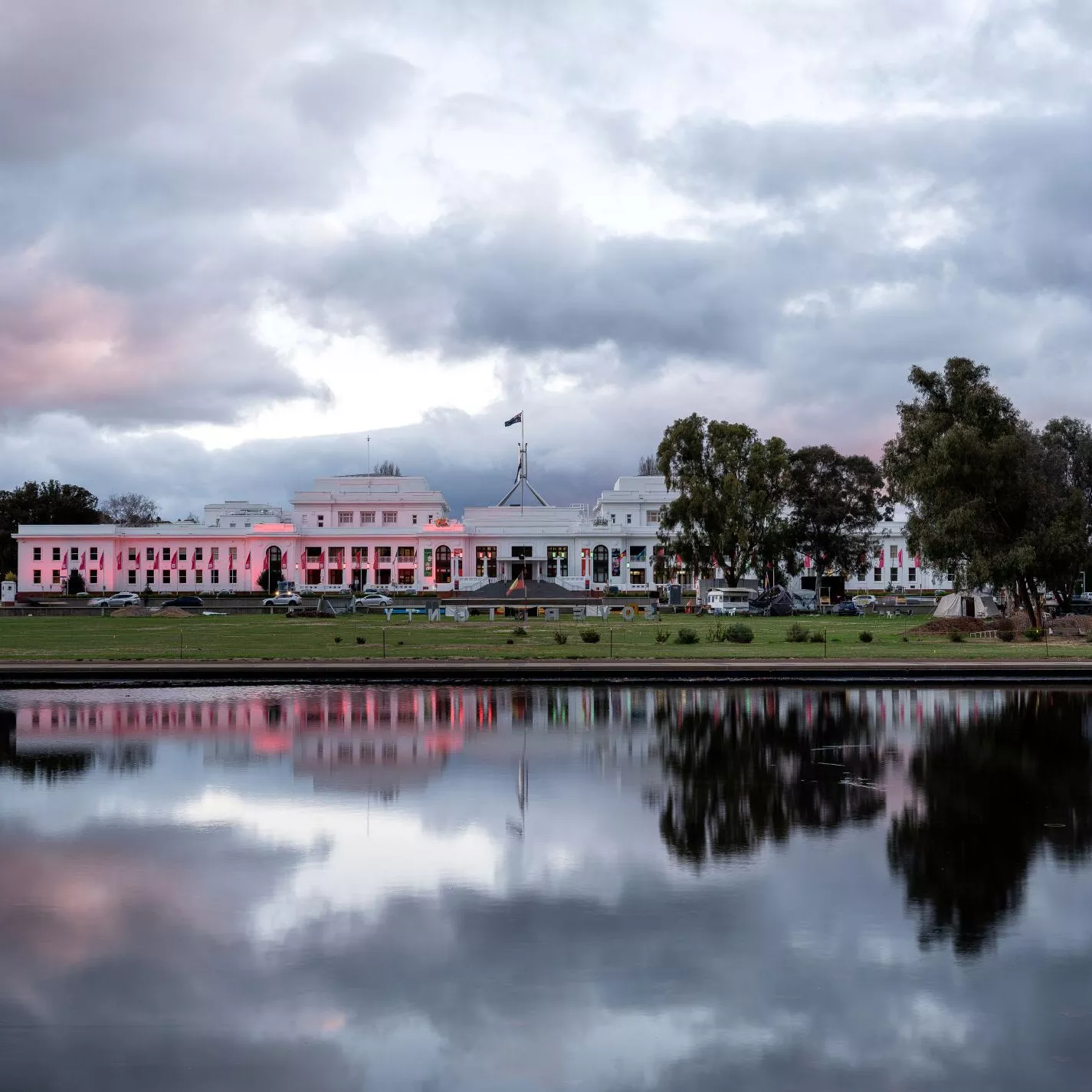 A large white building with the Australian flag on top, with a body of water in front that shows the reflection. The image is at dusk. 