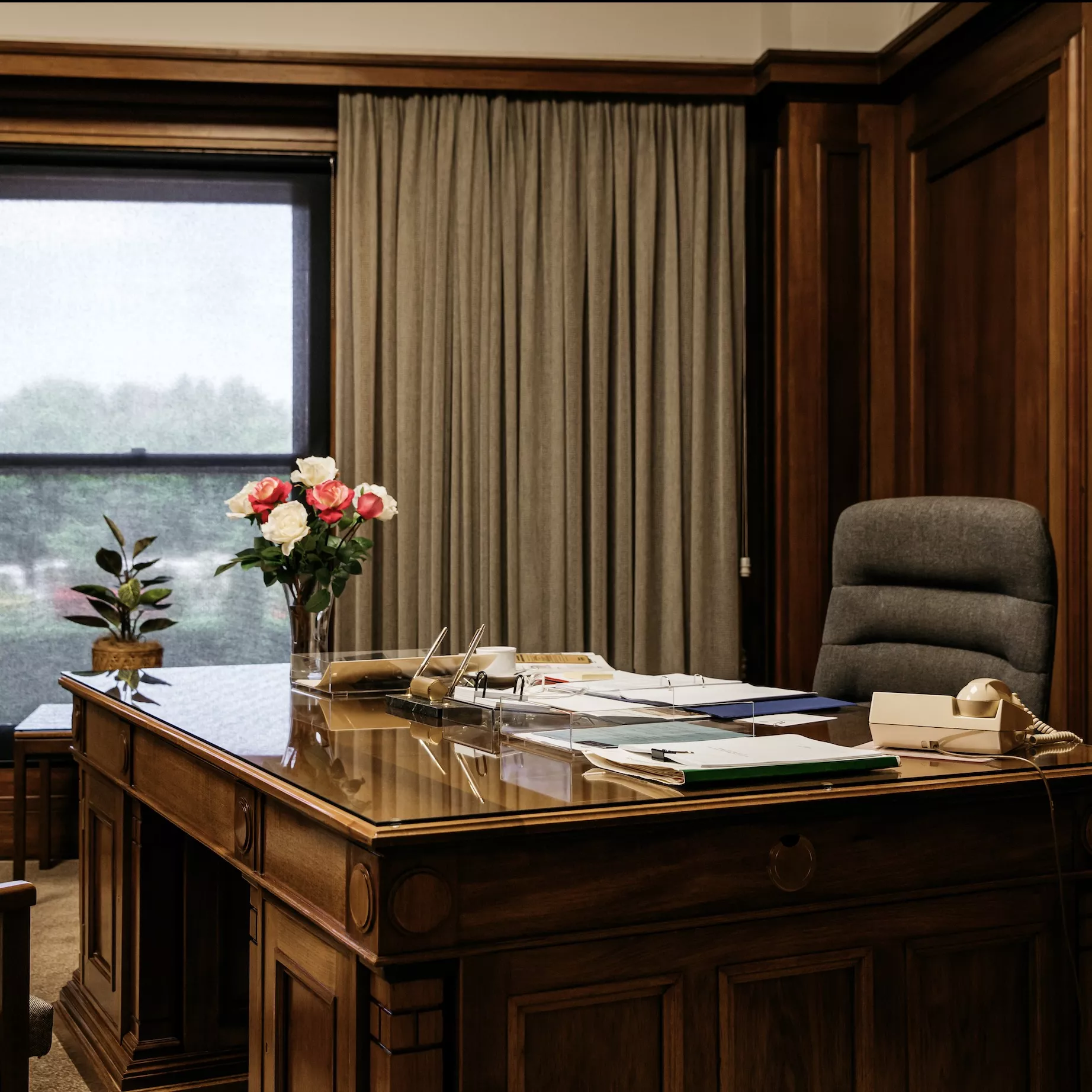 A large timber desk covered in folders and notes, with a vase with roses on it. 
