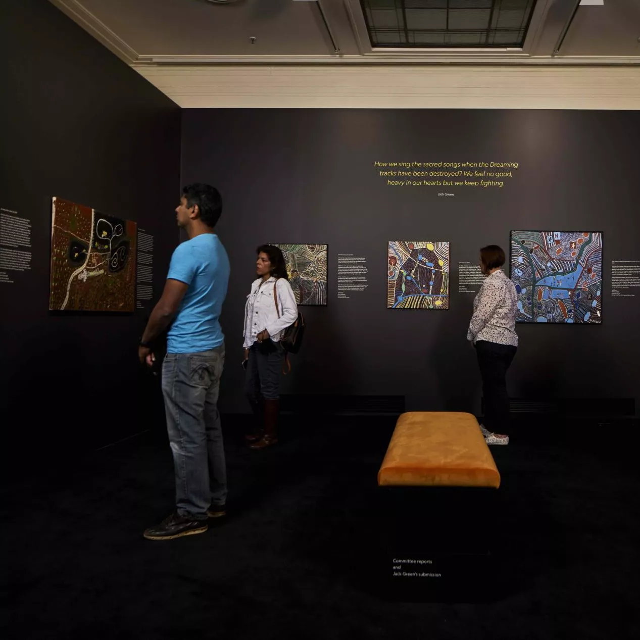 4 people stand in a museum room looking at paintings on the wall by artist Jack Green.