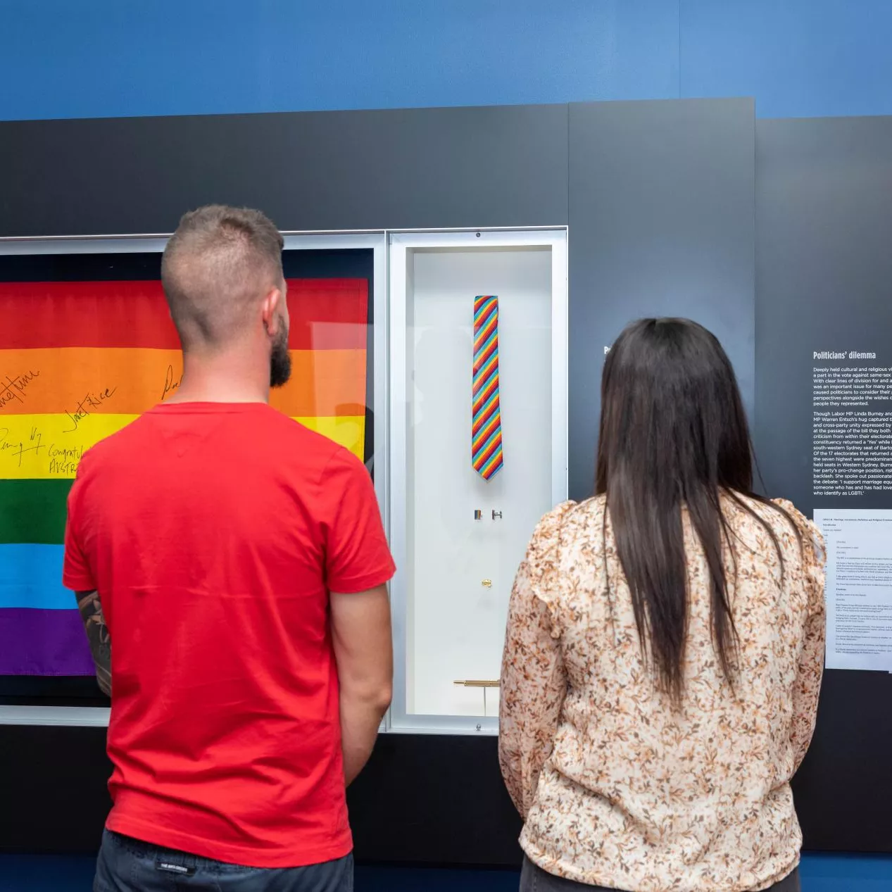 A man and a woman stand in front of a display case with a rainbow flag with signatures on it. 